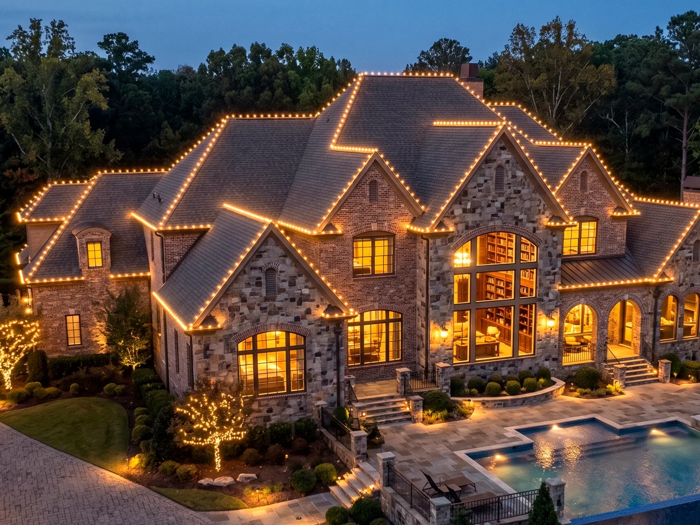 An aerial night view of a large stone and brick estate, every roofline and gable outlined in warm white lights, with a lit pool and patio below.